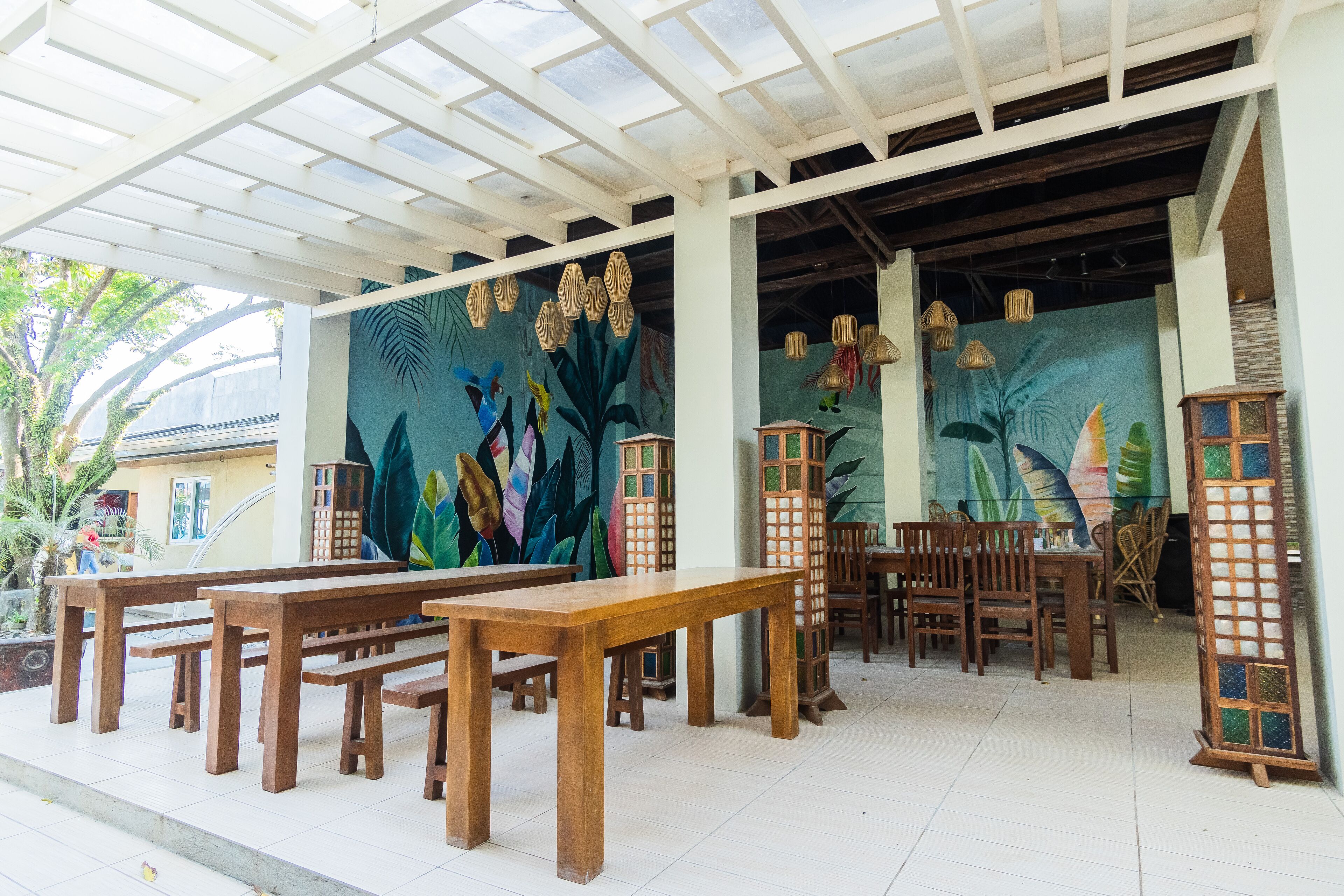 An outdoor seating area with wooden tables and benches, a mural with tropical leaves, and hanging wicker lamps.