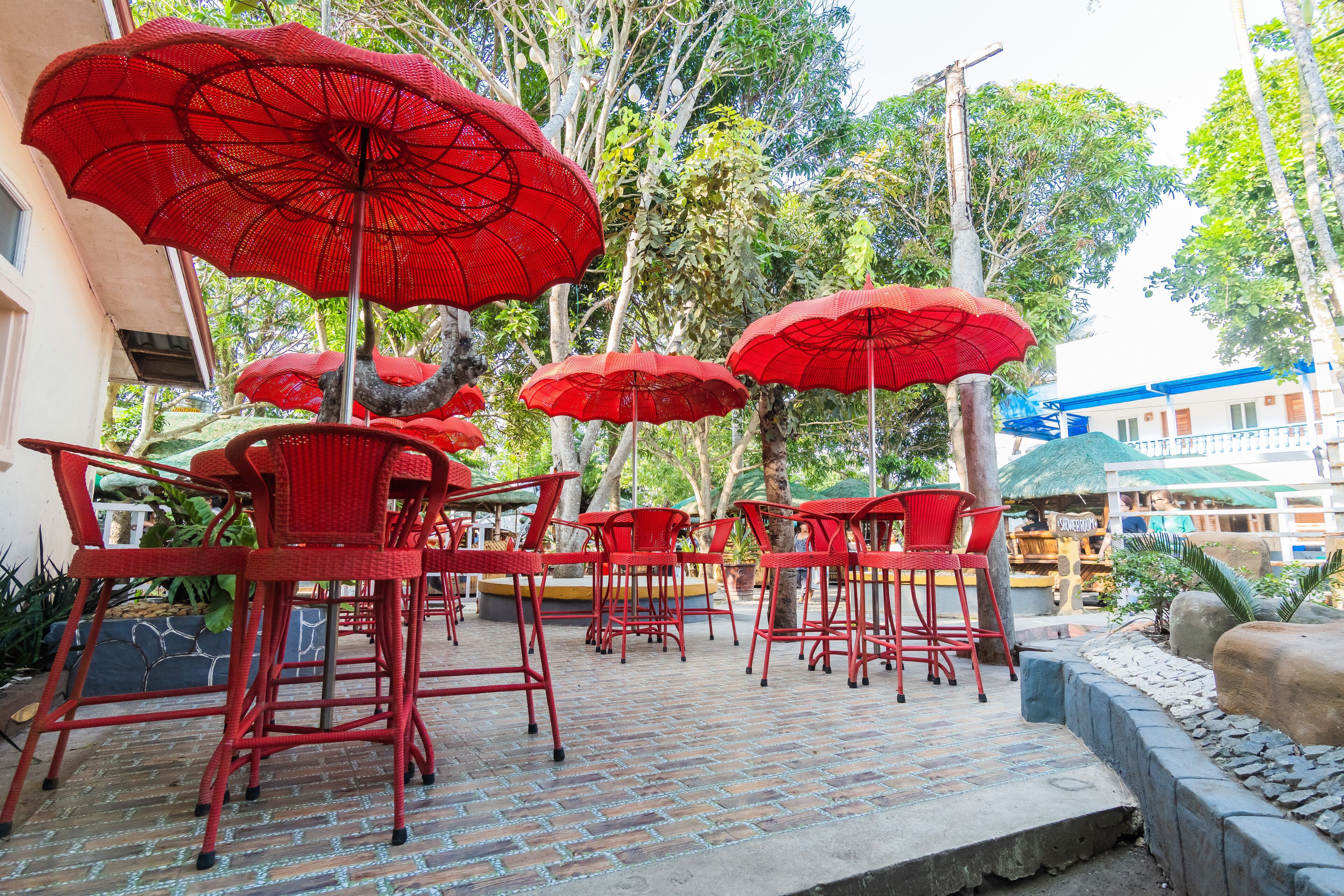 Outdoor seating with red chairs and umbrellas, surrounded by greenery and a building in the background.