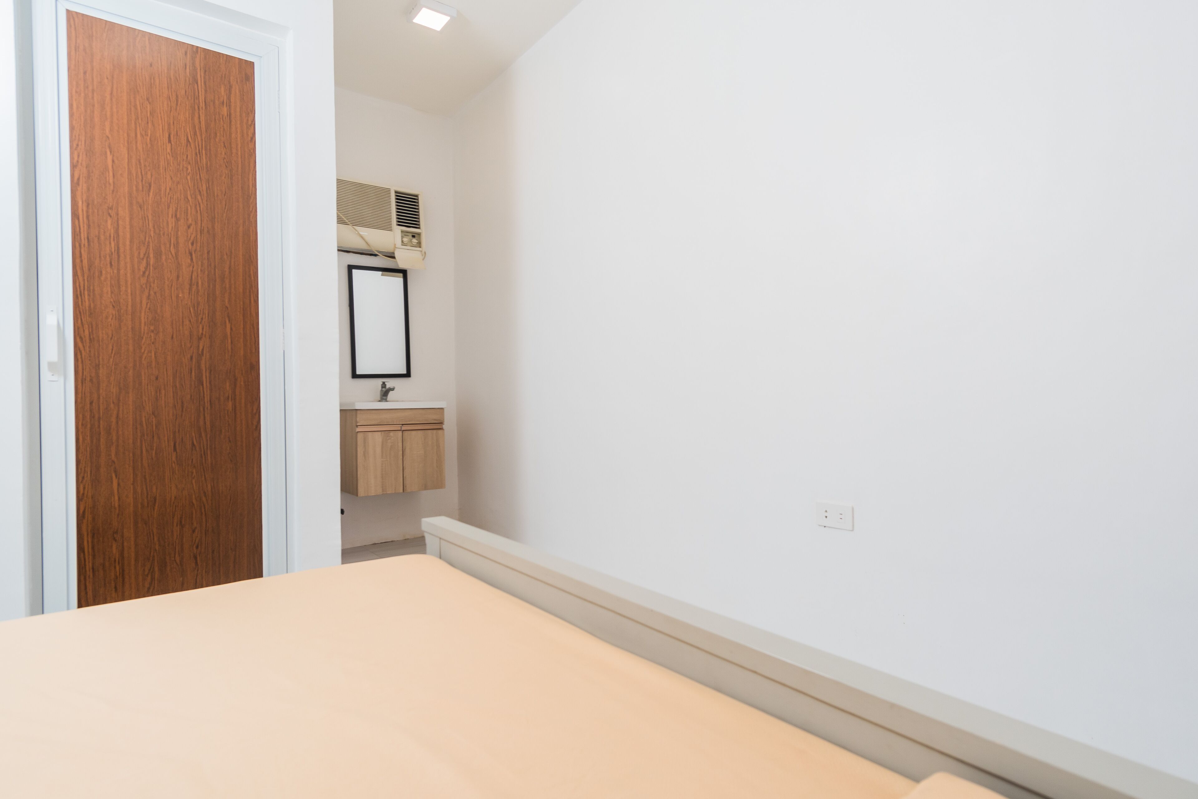 Une chambre avec une porte en bois, une petite salle de bain avec un miroir et un lavabo, et un mur blanc uni.