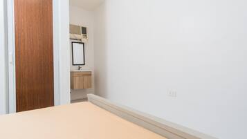 Une chambre avec une porte en bois, une petite salle de bain avec un miroir et un lavabo, et un mur blanc uni.