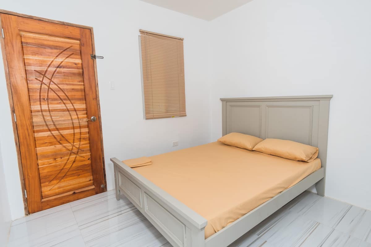 Ein Schlafzimmer mit Holztür, einem Bett mit beiger Matratze und einem Fenster mit Jalousien.