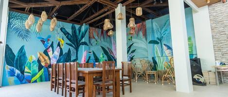 A dining area with wooden tables and chairs, a mural with tropical leaves, and hanging wicker lamps.