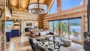 A spacious living area with a large window overlooking a lake, a fireplace, and a cozy seating area. - Lake Front! Mtn/Lake Views,HotTub,Gas FirePit,Walking Trail to town & shopping (Estes Park)
