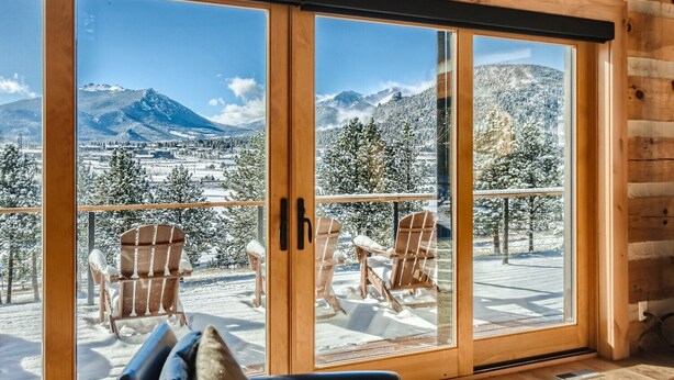 A wooden cabin with large sliding glass doors leading to a deck with two Adirondack chairs and a table. - Lake Front! Mtn/Lake Views,HotTub,Gas FirePit,Walking Trail to town & shopping (Estes Park)