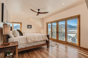 Une chambre avec un lit, un ventilateur de plafond, une grande fenêtre donnant sur des arbres et des montagnes enneigés, et un plancher en bois.