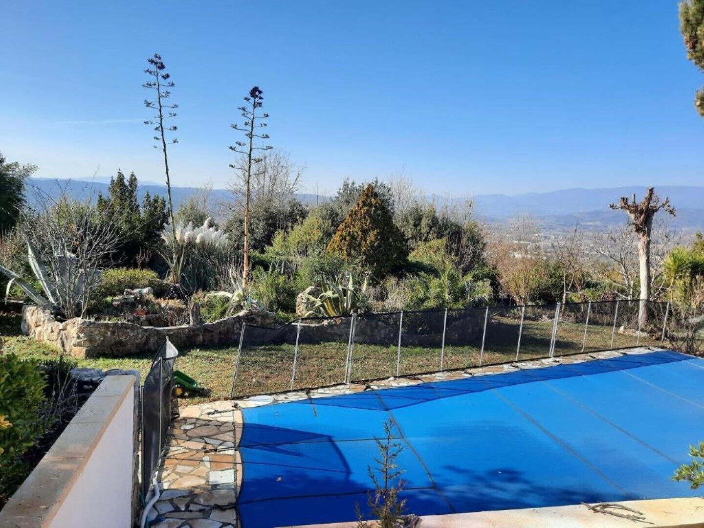 A swimming pool with a blue cover, surrounded by a stone wall and various plants.