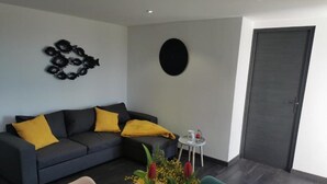 Una sala moderna con un sofá gris oscuro, cojines amarillos, un adorno negro redondo en la pared, una pequeña mesa redonda con un objeto rojo y una mesa de comedor de madera con sillas.