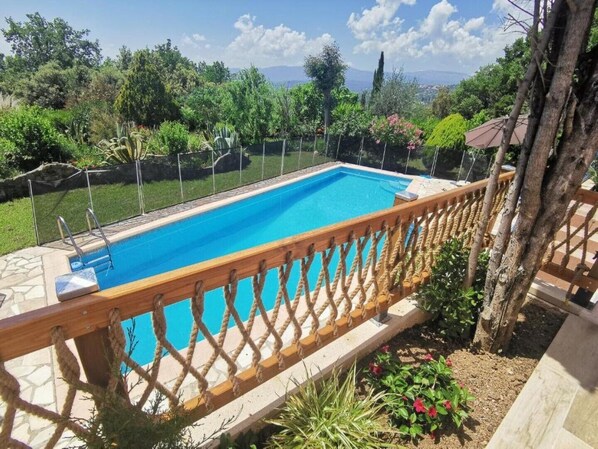 Ein rechteckiger Pool mit Holzterrasse und Seilgeländer, umgeben von üppiger Vegetation und blauem Himmel.