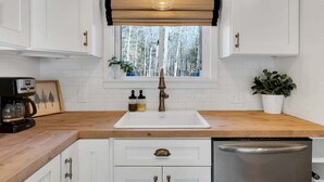 A kitchen with white cabinets, a wooden countertop, a stainless steel dishwasher, a window with a view of trees, and a hanging light fixture.