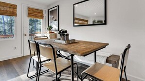 A dining area with a wooden table, four chairs, a large mirror, and a view of the outdoors through a window with a bamboo blind.