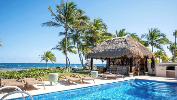 A pool area with lounge chairs, a thatched-roof structure, and palm trees.