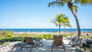 Two wicker lounge chairs facing a beach with a palm tree and ocean view.