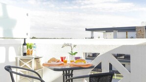 A balcony with a table set for breakfast, including croissants, orange juice, and a bottle of wine, overlooking a modern building and a body of water. - Casa Andorinhas - Quinta Velha Village - Free WiFi and Meo (Cabanas de Tavira)