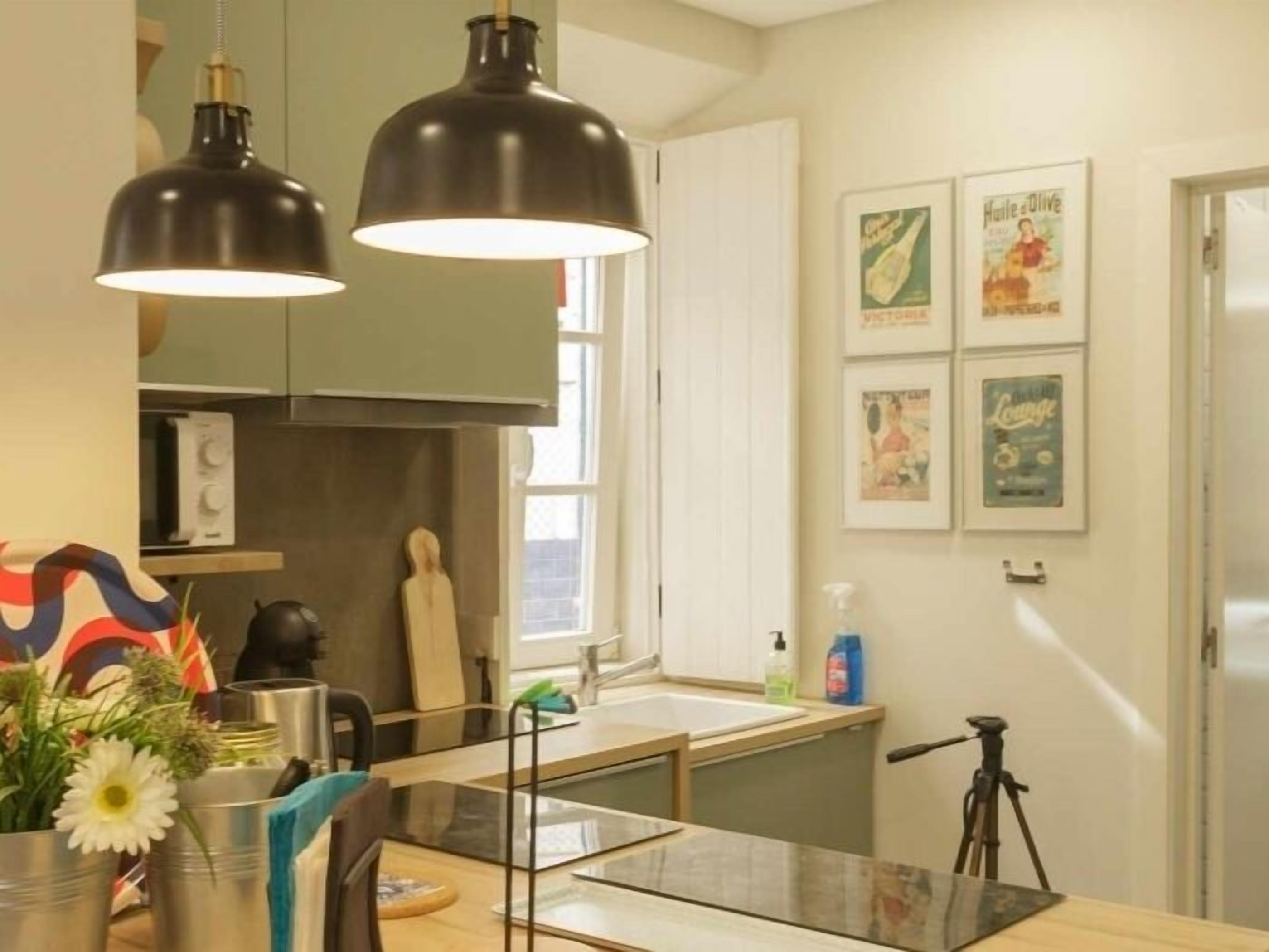 A modern kitchen with two pendant lights, a sink, and a window. There are framed pictures on the wall and a tripod in the corner.