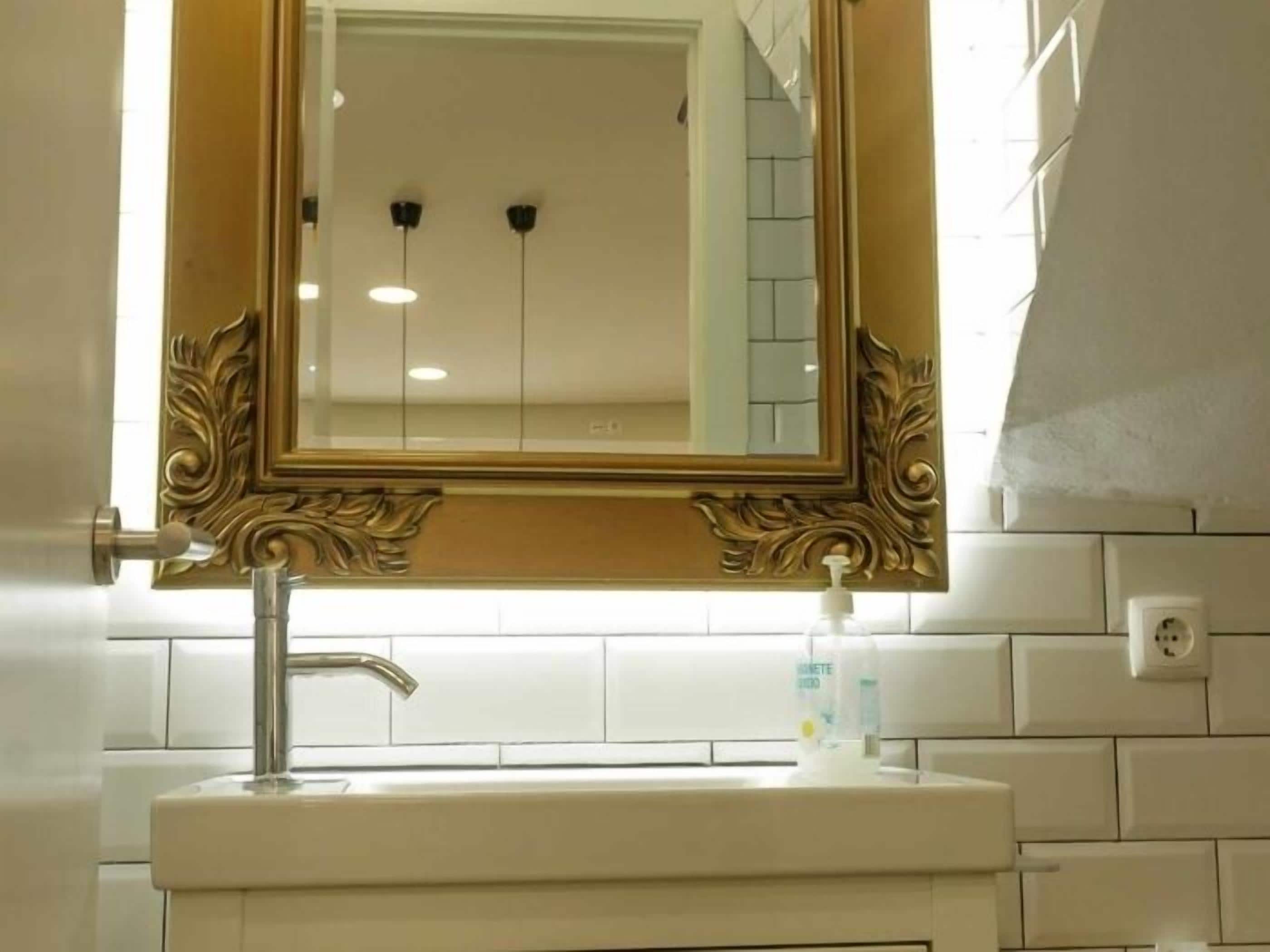 A bathroom with a white sink, a gold-framed mirror, and a white tiled wall.