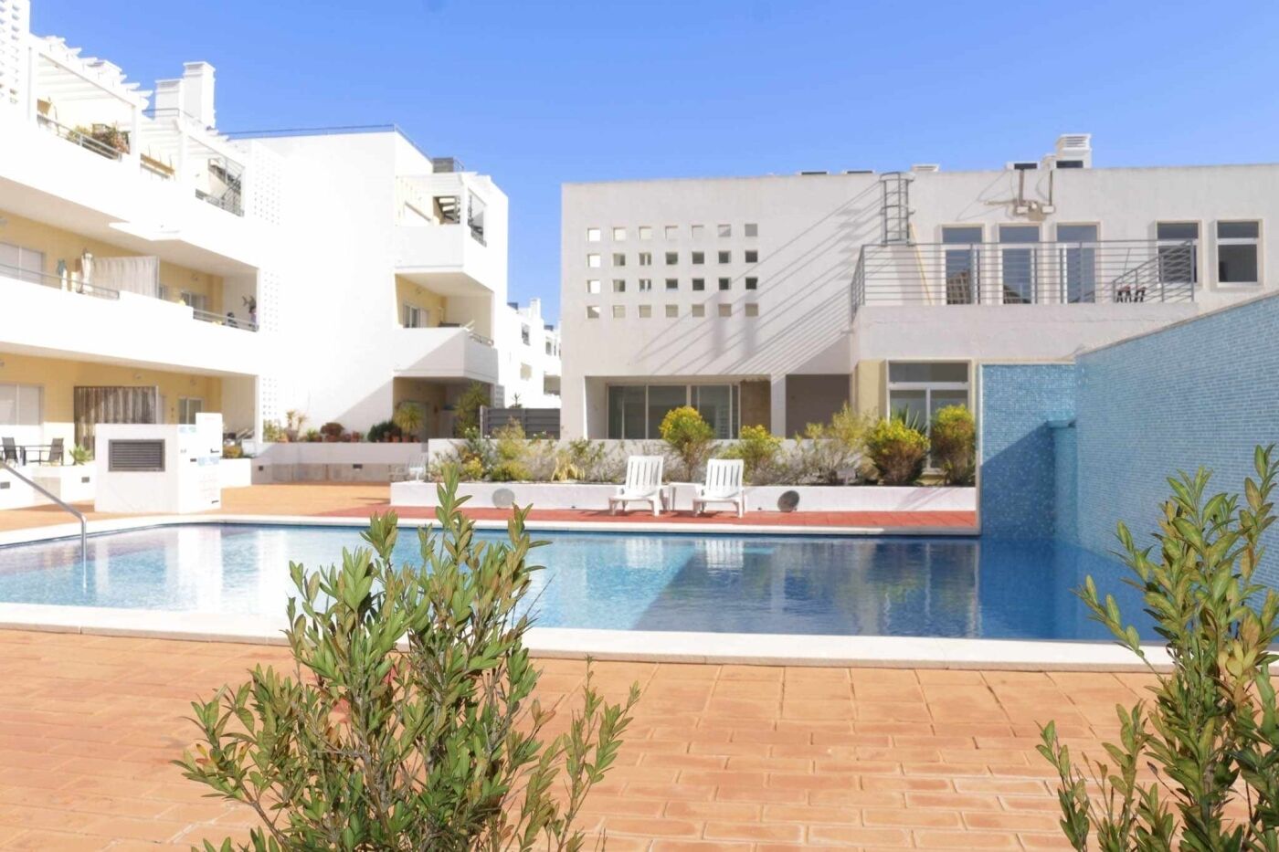 A swimming pool with lounge chairs and a brick patio, surrounded by modern buildings with balconies and windows.