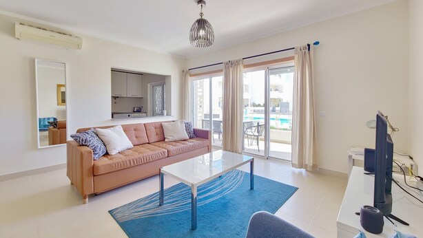 Ein modernes Wohnzimmer mit Sofa, Couchtisch und Fernseher. Im Hintergrund befindet sich eine Küchenzeile und durch die Schiebetüren ist ein Schwimmbecken zu sehen.
