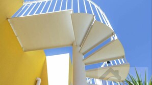 Eine Wendeltreppe mit weißer Brüstung und beigen Stufen führt zu einem Balkon mit einer gelben Wand und blauem Himmel im Hintergrund.