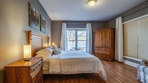 Une chambre avec un plancher en bois, un lit avec une literie blanche, une table de chevet avec une lampe, une commode en bois et une fenêtre avec des rideaux.