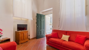 Un salon avec un canapé rouge, un téléviseur posé sur un meuble en bois et une porte donnant sur l’extérieur.
