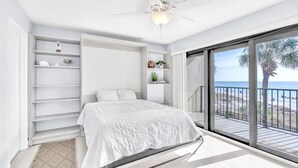 Una camera da letto con un letto, scaffali bianchi, un ventilatore a soffitto e un balcone vista spiaggia.