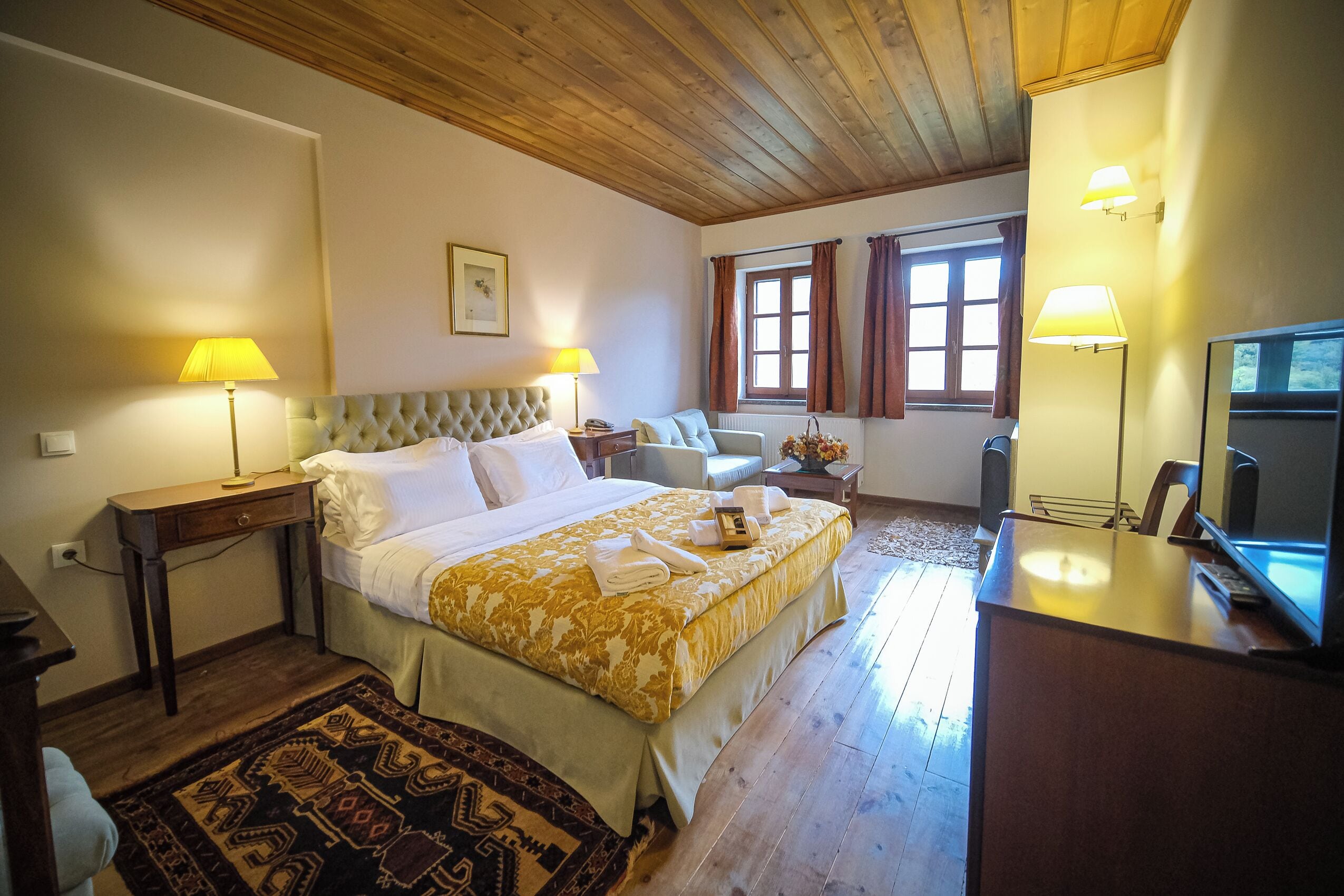 A hotel room with a large bed, two bedside lamps, a wooden ceiling, a desk with a television, and a window with curtains.