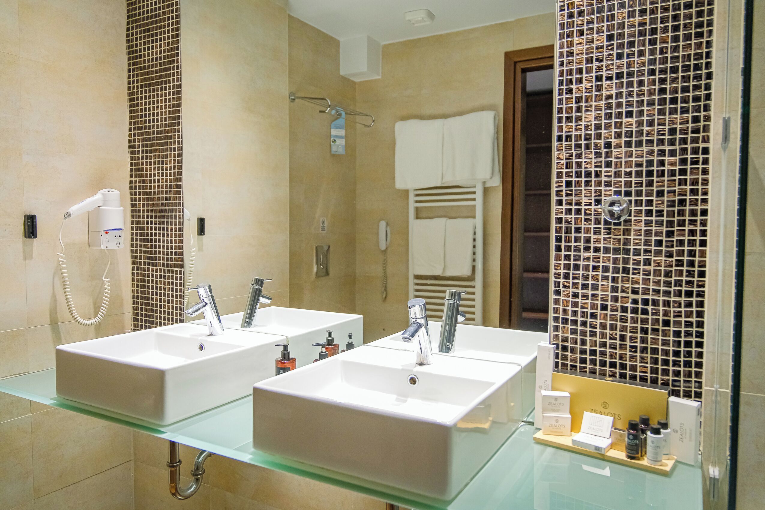 A bathroom with two sinks, a hairdryer, and a glass shelf with toiletries.