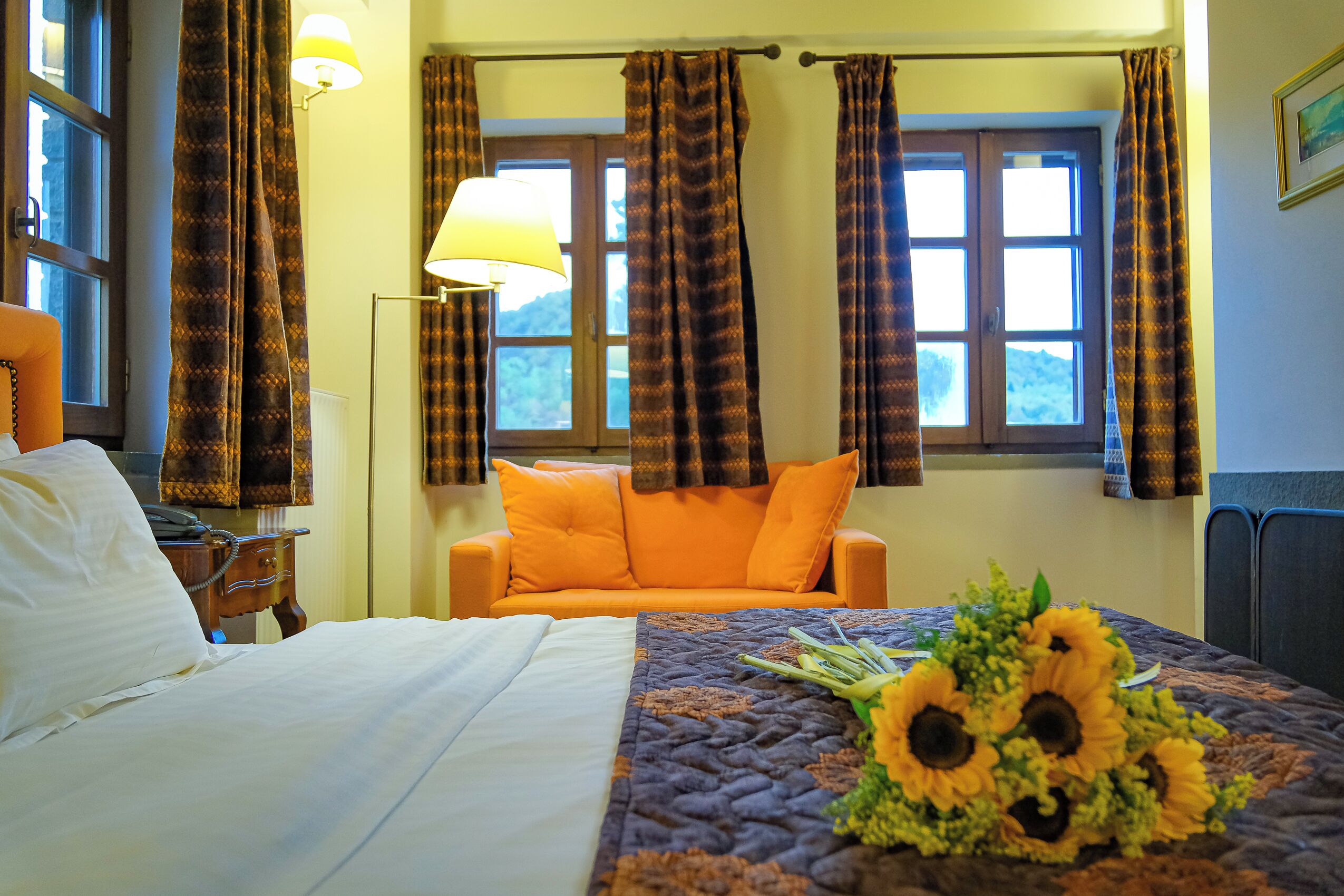 Een hotelkamer met een bed, een oranje bank, een raam met gordijnen, een lamp en een bos zonnebloemen op het bed.