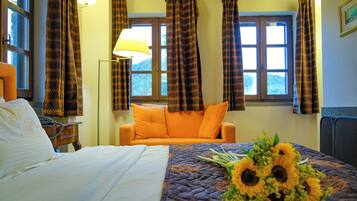 Habitación de hotel con cama, sofá naranja, ventana con cortinas, lámpara y un ramo de girasoles sobre la cama.