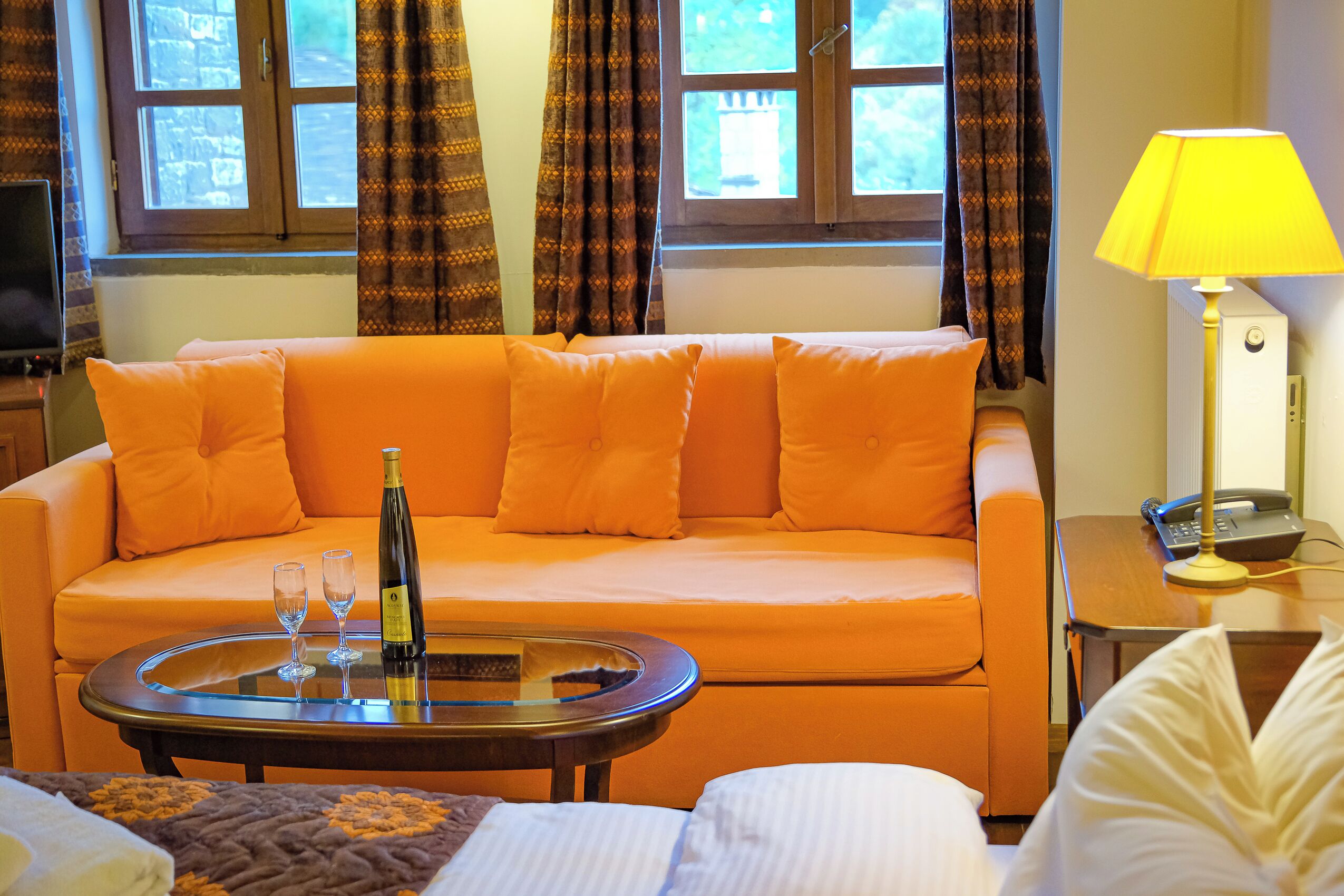A room with a sofa, a table with a bottle and glasses, a lamp, and a bed.