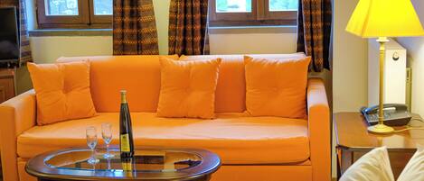A room with a sofa, a table with a bottle and glasses, a lamp, and a bed.