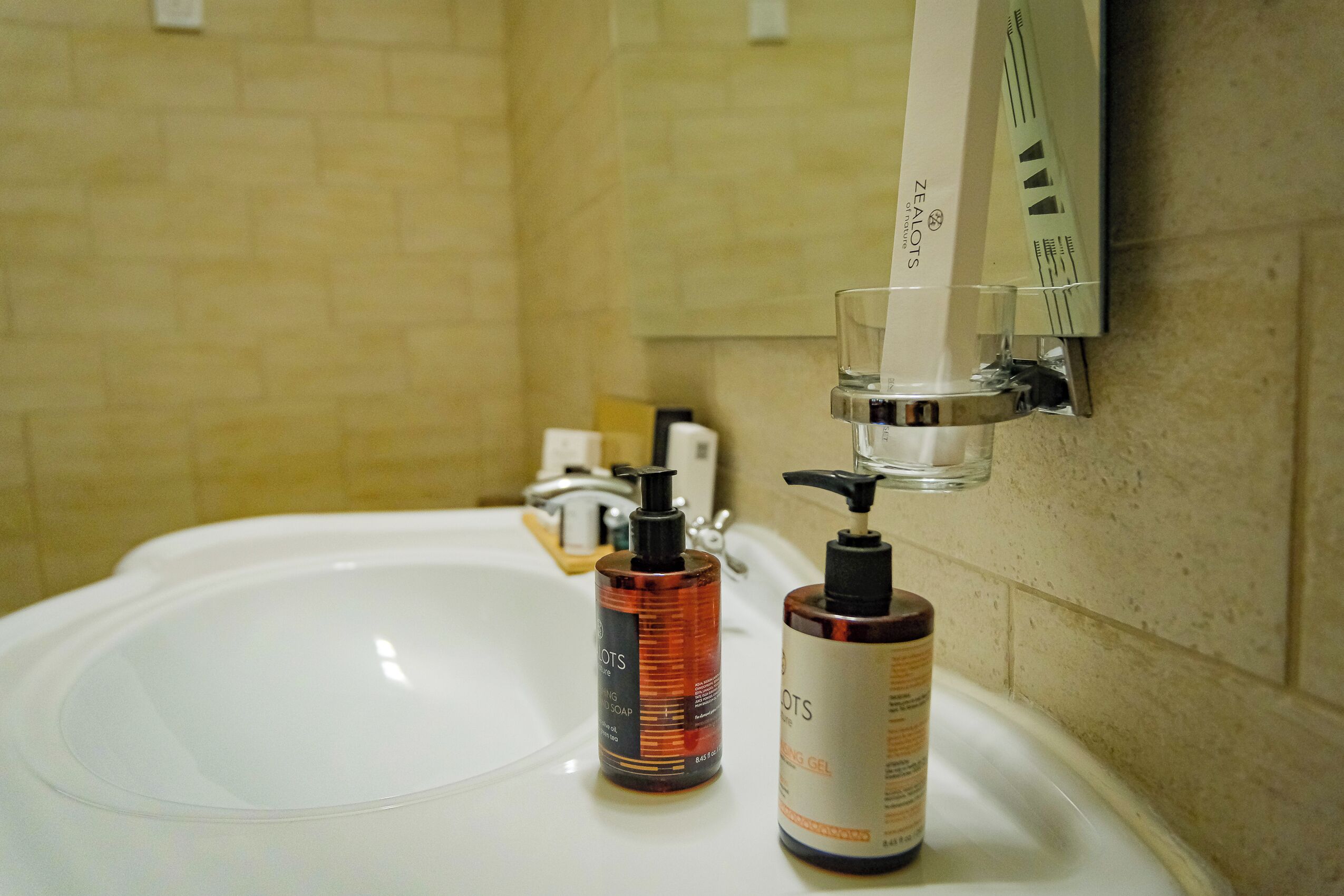 A bathroom sink with two bottles of Zealots products, a soap dispenser, and a glass of water.