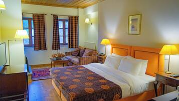 Quarto de hotel com uma cama grande, duas mesas de cabeceira com luminárias, uma cadeira, uma escrivaninha e uma janela com cortinas.