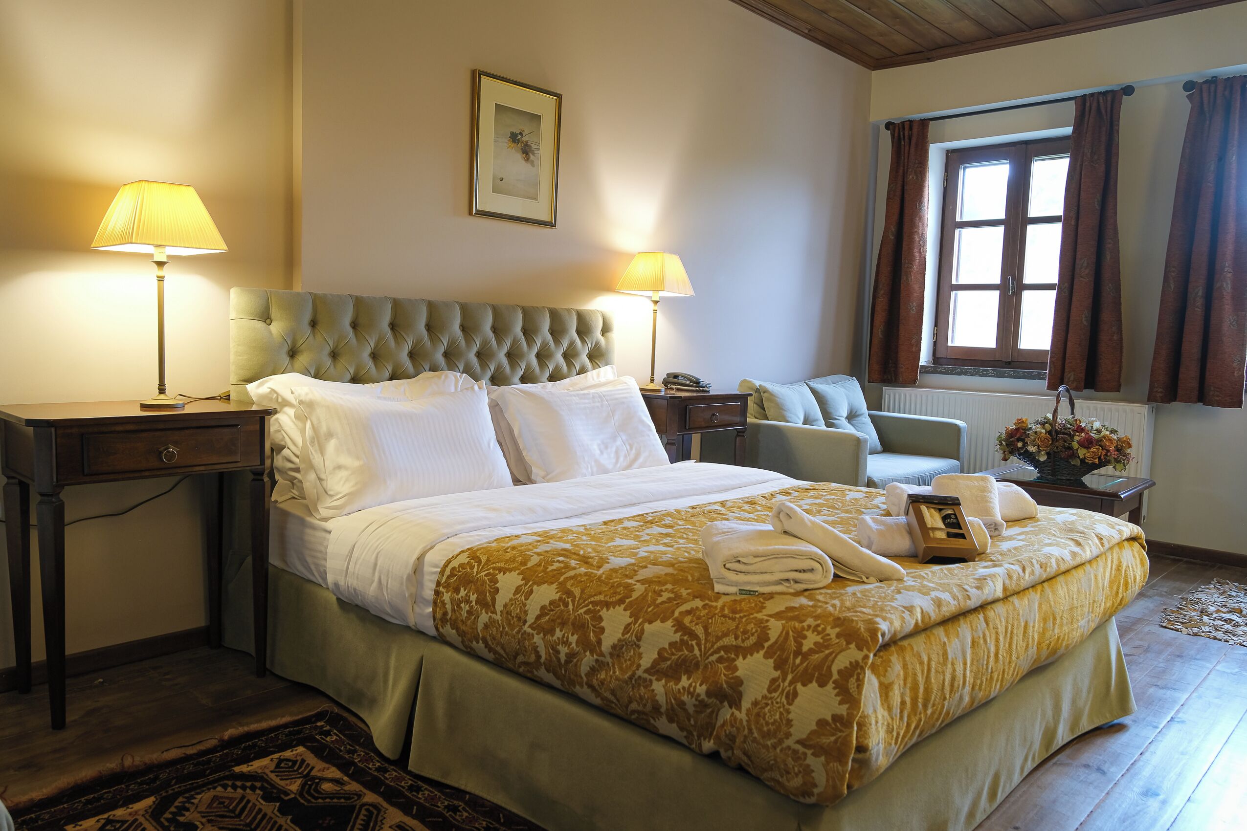 A neatly made bed with a floral bedspread, two bedside lamps, a bedside table with a phone, and a window with curtains.