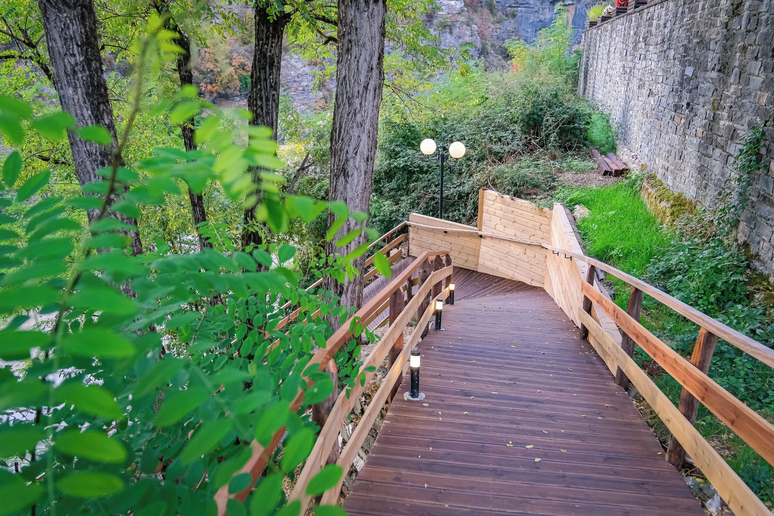 Een houten brug met leuningen en lantaarns, omringd door groen en een stenen muur.