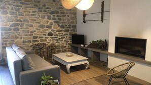 Uma sala de estar com parede de pedra, um sofá cinzento, uma mesa de centro branca, uma poltrona de vime e uma lareira.