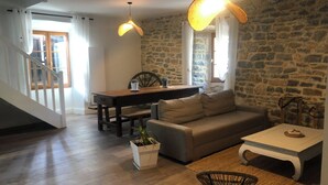 Uma sala de estar com parede de pedra, um sofá, uma mesa de centro, uma mesa de jantar com cadeiras, uma escada e um vaso com planta.