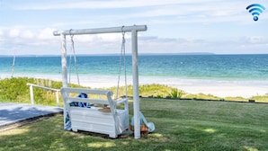 A white swing chair with a basket and a bag, overlooking a beach with clear blue water and a sandy shore.