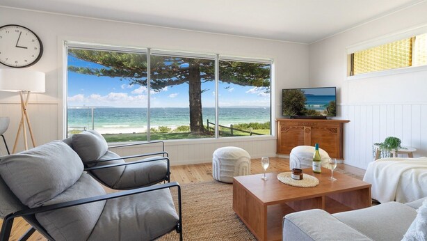 A room with a large window overlooking a beach, a wooden table with a bottle and glasses, a sofa, and a clock on the wall.