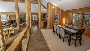 Uma sala de estar ampla com pé direito alto, vigas de madeira, mesa de jantar com cadeiras e grandes janelas que oferecem vista para o exterior.