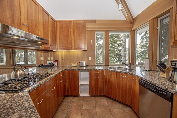 Una cocina con armarios de madera, encimeras de granito, electrodomésticos de acero inoxidable y grandes ventanas que dan a vistas de árboles.