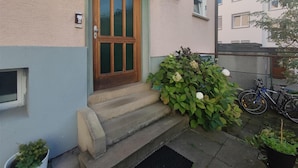 A residential building with a wooden door, steps leading to the entrance, and a small garden with white flowers.