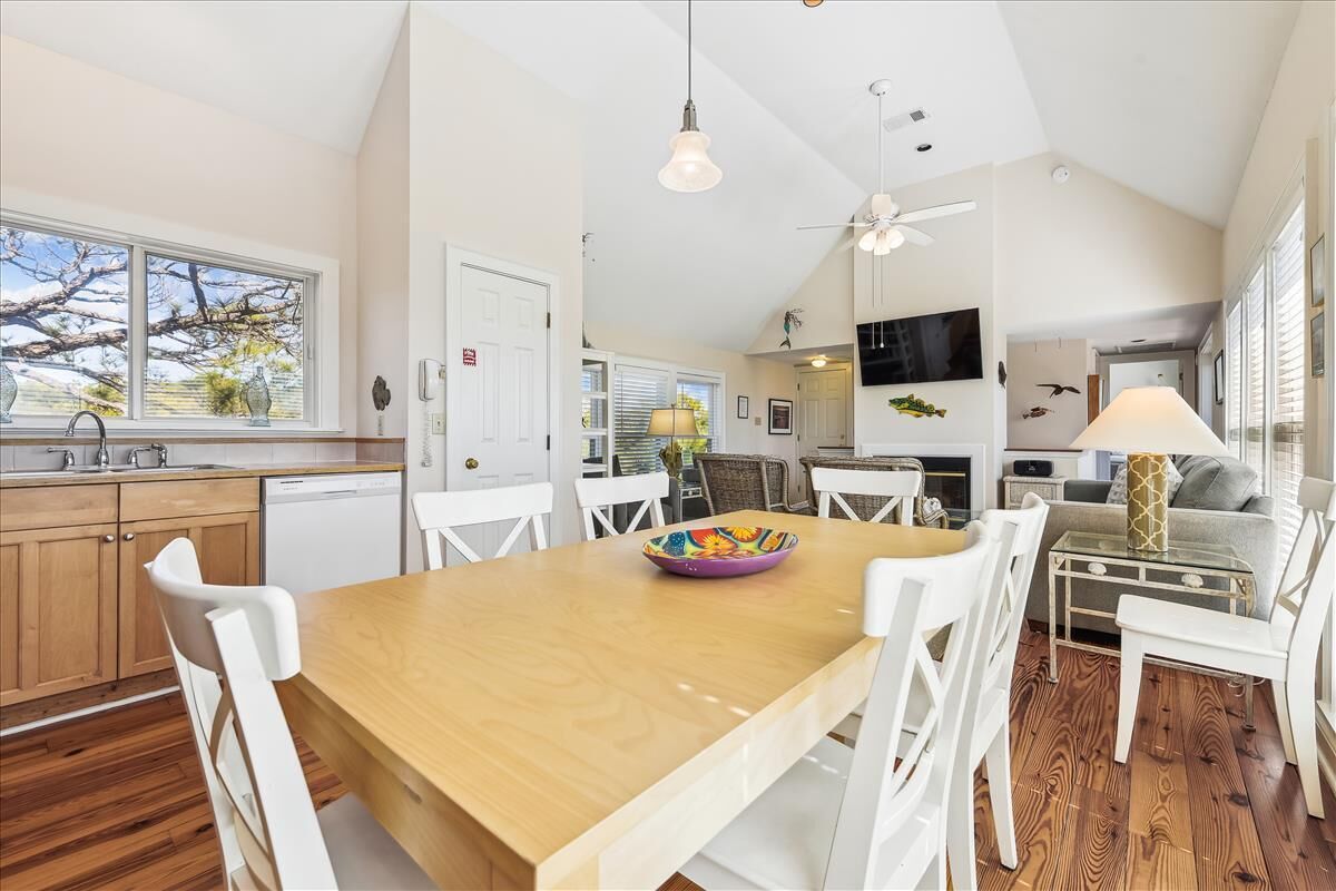 A spacious kitchen with a wooden table, white chairs, and a colorful bowl. There is a dining area with a sofa and a TV mounted on the wall.