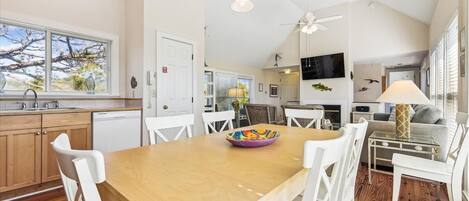 A spacious kitchen with a wooden table, white chairs, and a colorful bowl. There is a dining area with a sofa and a TV mounted on the wall.