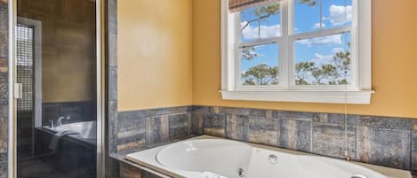 Une salle de bain dotée d’une grande baignoire, d’un espace douche et d’une fenêtre offrant une vue sur les arbres et le ciel.