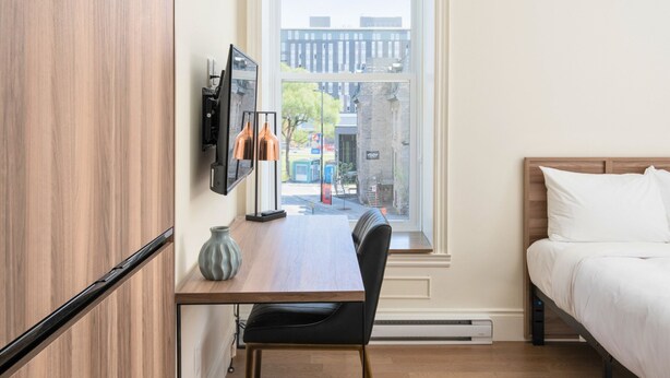 A modern hotel room with a wooden floor, a bed with white bedding, a desk with a chair, a mounted TV, and a window with a city view. - Opal Lofts - Quartier Latin (Montreal)