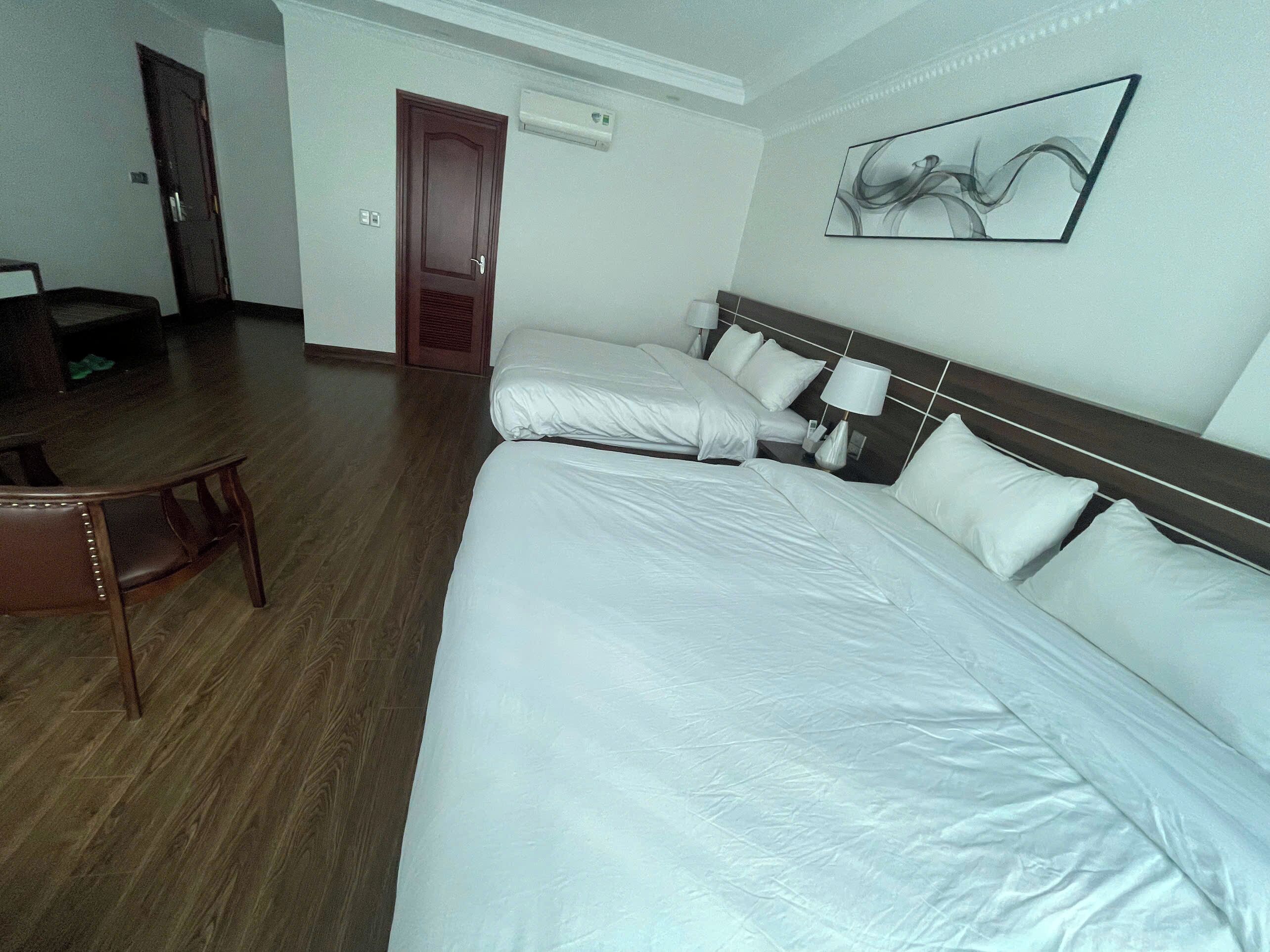 A hotel room with two beds, a wooden floor, a door, and a wall-mounted air conditioner.