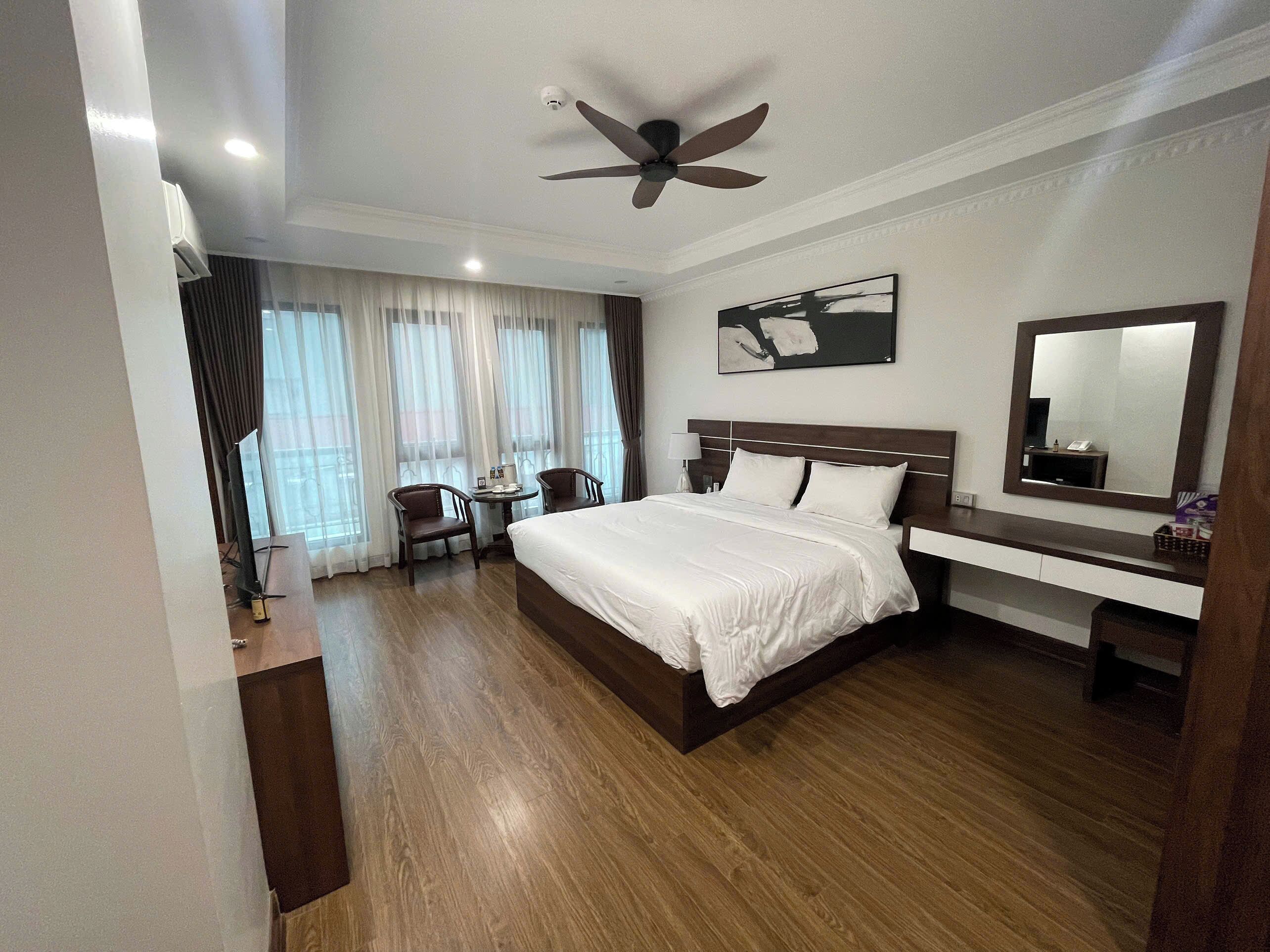 A hotel room with a large bed, a desk with two chairs, a ceiling fan, a large mirror, and a window with curtains.