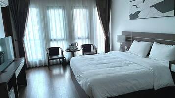 Habitación de hotel con una cama grande, un escritorio con televisor, dos sillones, una mesita de noche con lámpara y una ventana con cortinas.