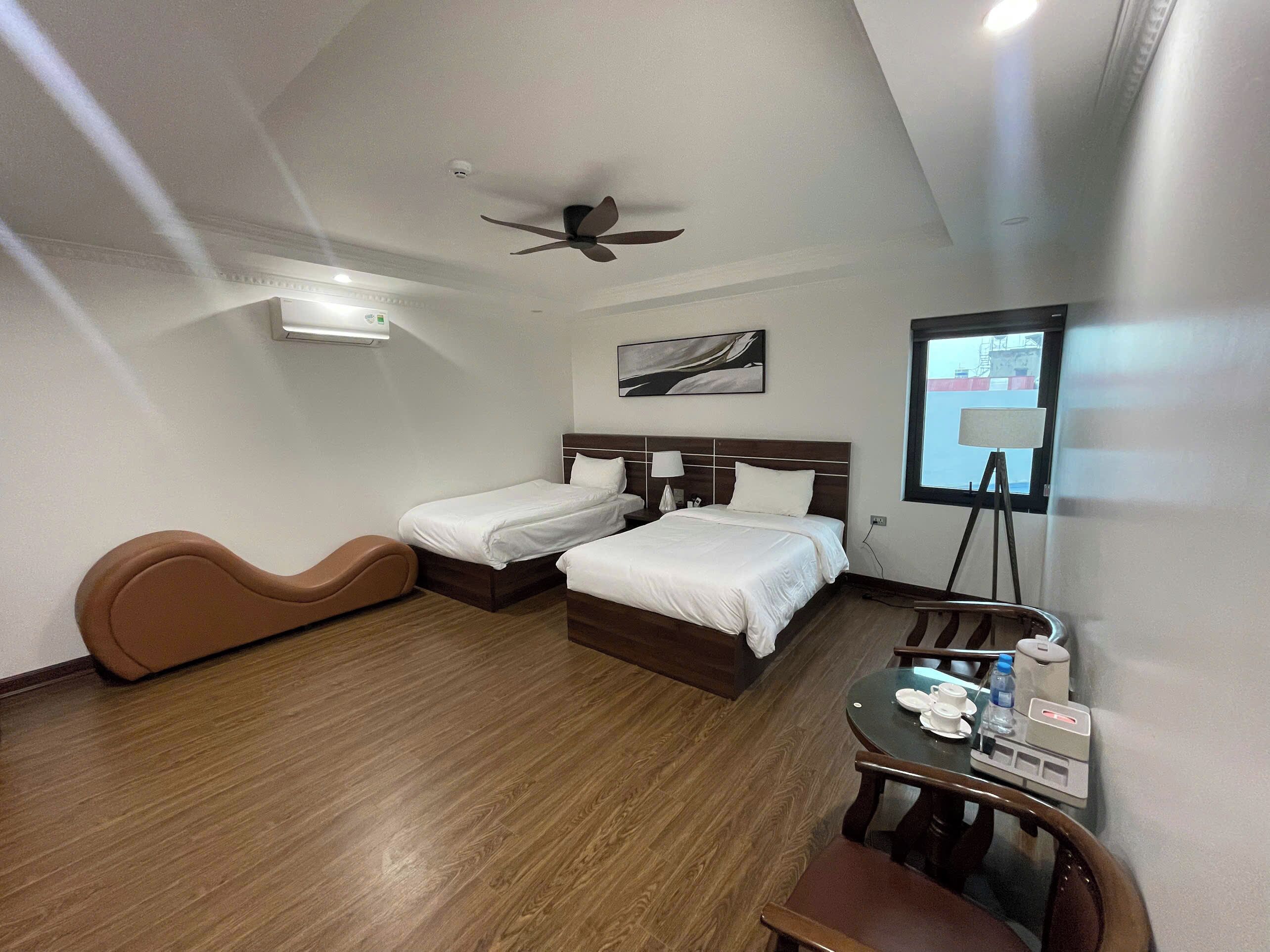 Una habitación de hotel con dos camas, un ventilador de techo, un escritorio con silla y una ventana con vistas.